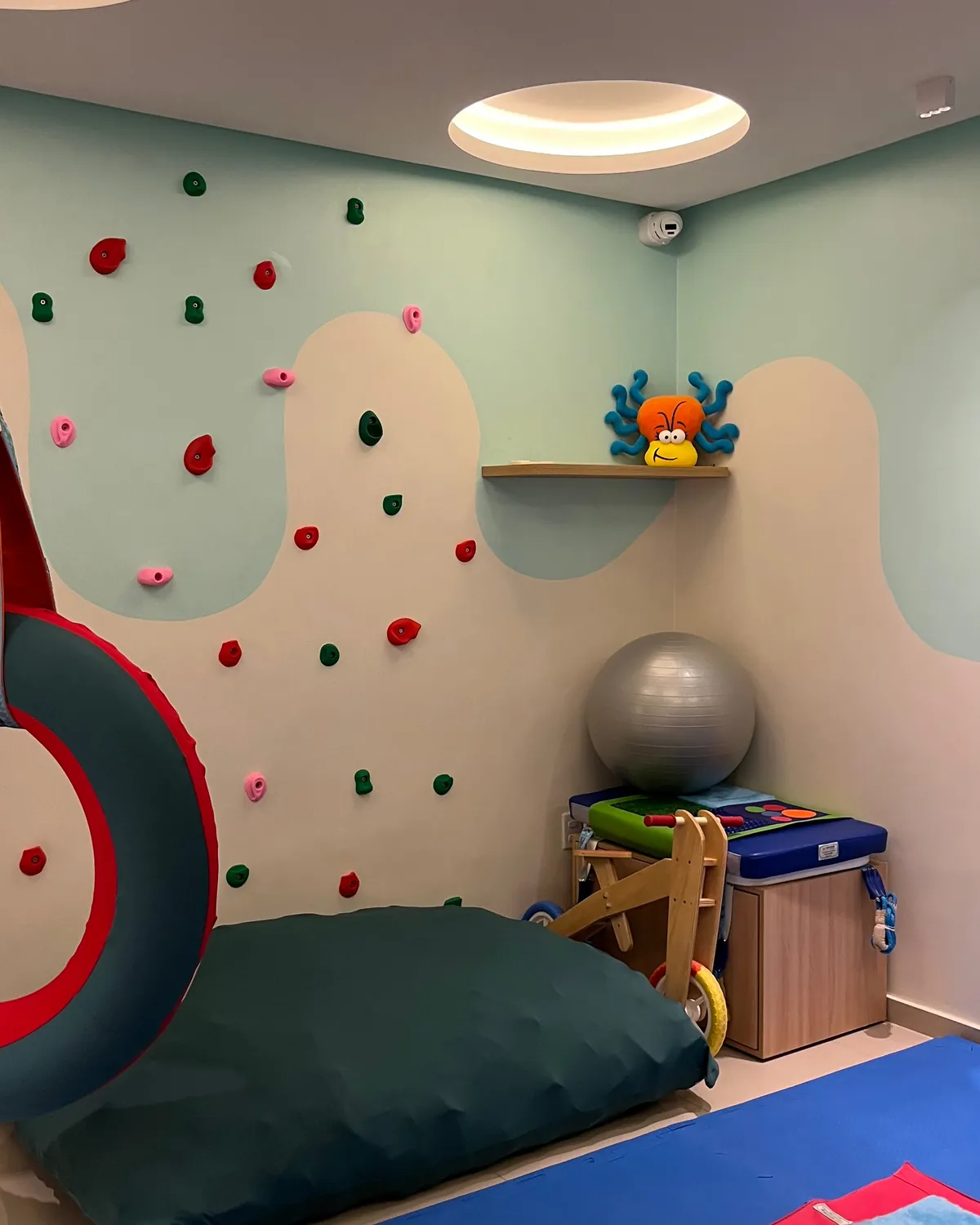 Canto da sala sensorial com almofadão verde, tatame azul, bola cinza e parede de escalada