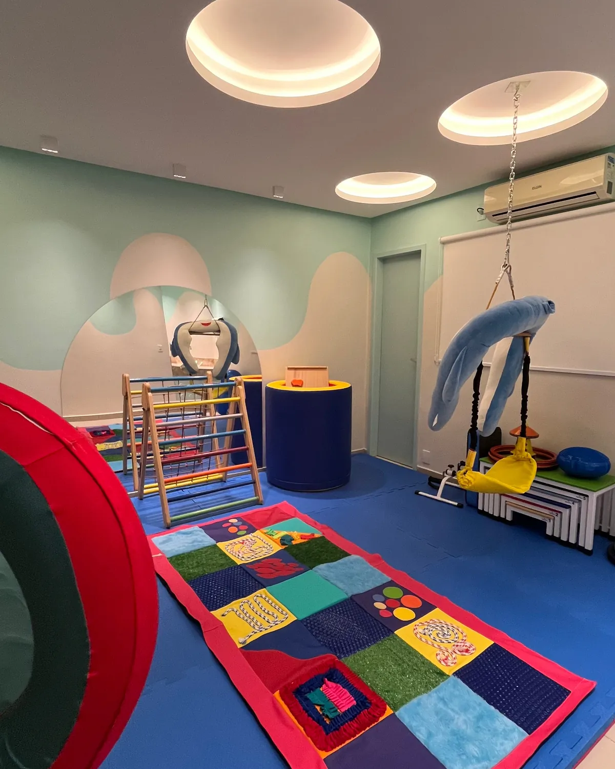 Sala sensorial com tatame azul e colorido, triângulo de escalada e balanços