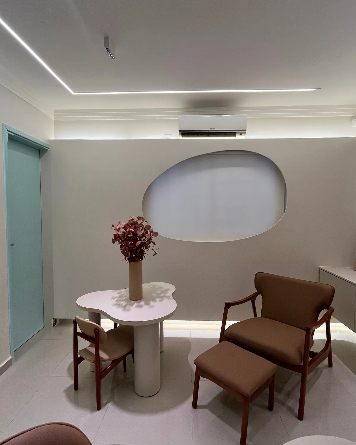 Poltrona com apoio para os pés ao lado de uma mesinha com flores, perto da janela oval do consultório