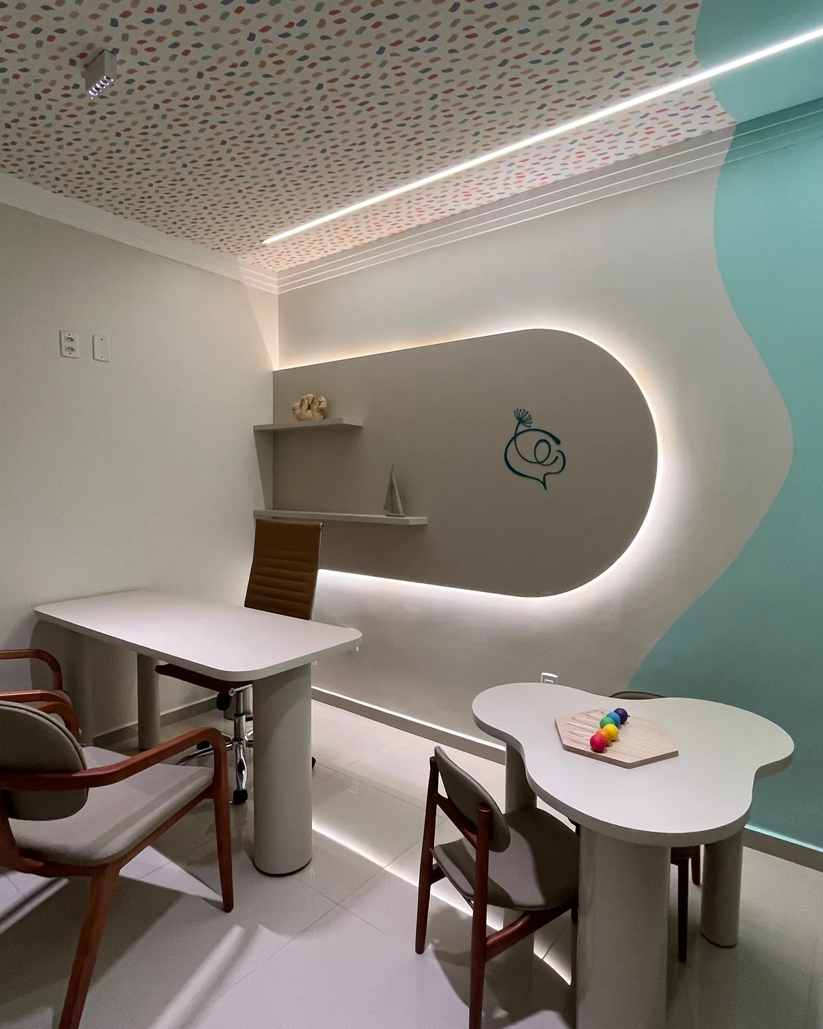 Consultório infantil com mesinha em forma de nuvem, um jogo colorido sobre a mesa, cadeiras e parede iluminada