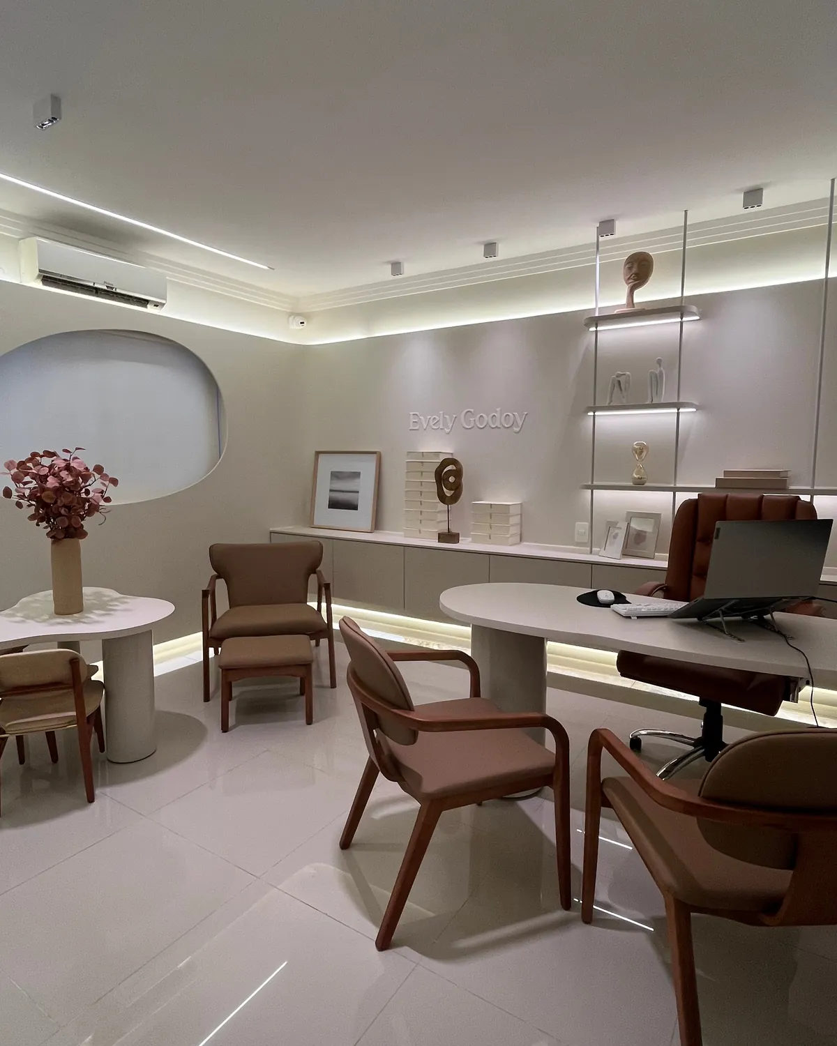 Consultório adulto com mesa redonda e flores, poltrona, mesa de atendimento e estante iluminada