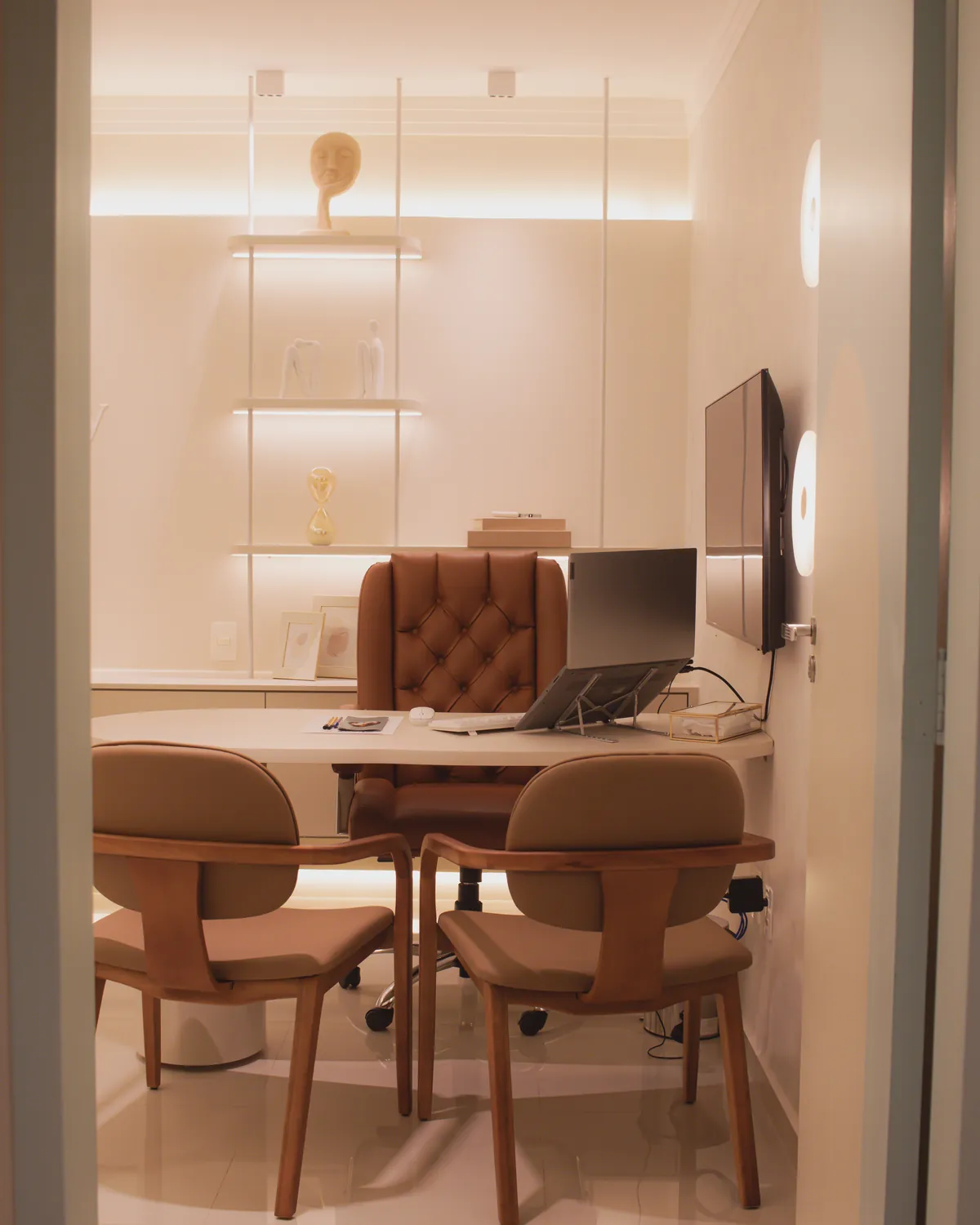 Consultório adulto visto da porta, com mesa de atendimento, cadeiras e estante iluminada